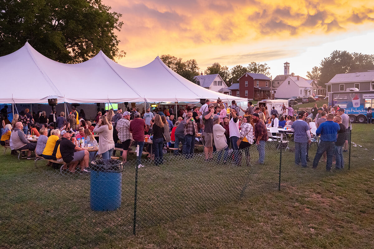 Crowd of people sit at picnic tables or stand in groups under a large white tent inside a temporarily fenced area. Historic German buildings, including a clock tower, are in the background and the clouds glow with dim light from a setting sun.